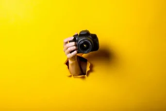 تصویر دست زنی که دوربینی را در پس زمینه زرد روشن در دست گرفته است